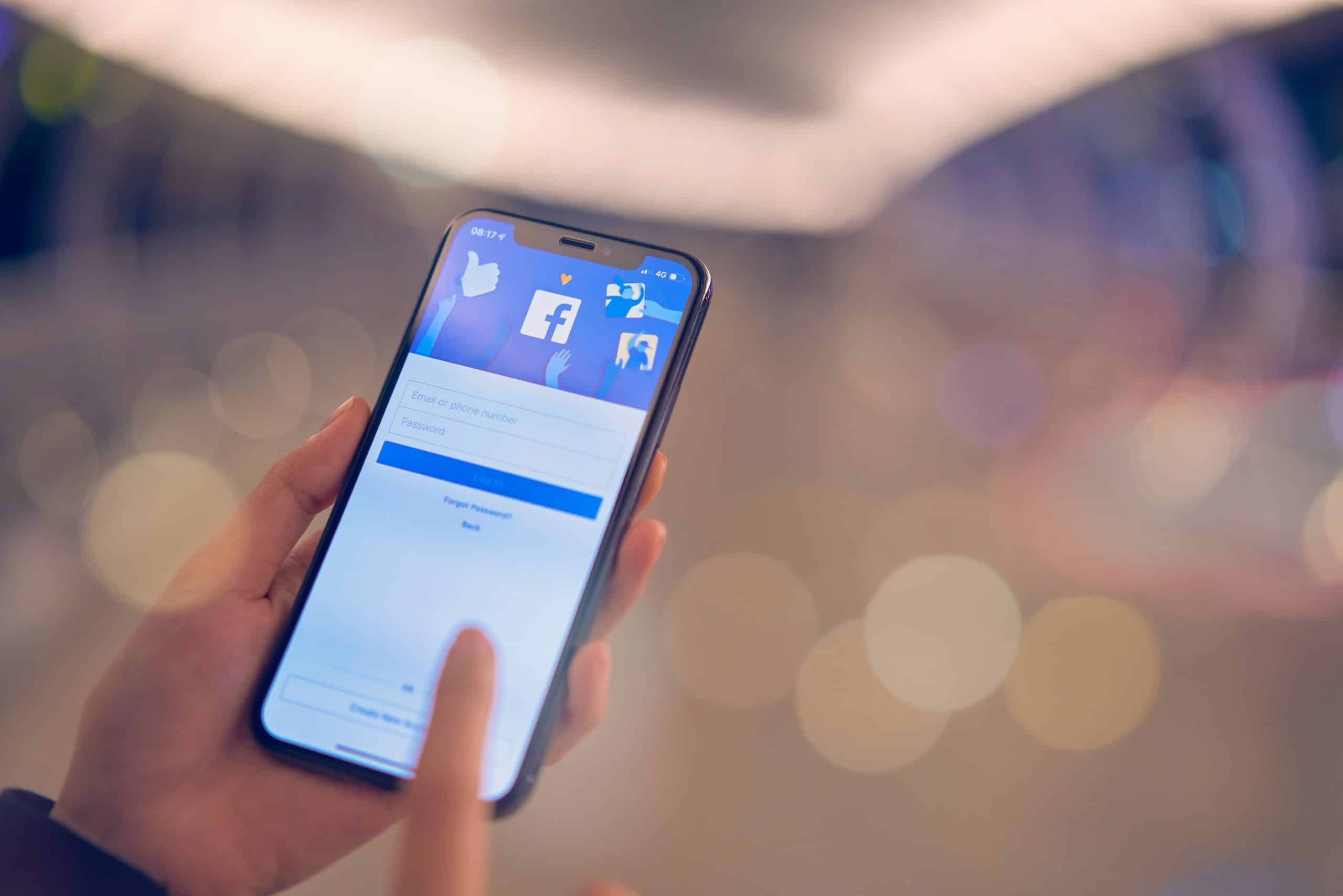 Close-up of a person's hand holding a smartphone with the Facebook login screen displayed. The person is tapping the screen, taking advantage of iOS 14's feature to view apps without downloading, while the background is blurred with bokeh lights.