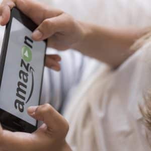 Close-up of a person holding a smartphone displaying the Amazon Prime logo with the play button, likely indicating a streaming service. The person appears to be lying down on a bed, wearing a light-colored shirt and striped pants, ready to rent or buy content for their next movie night.