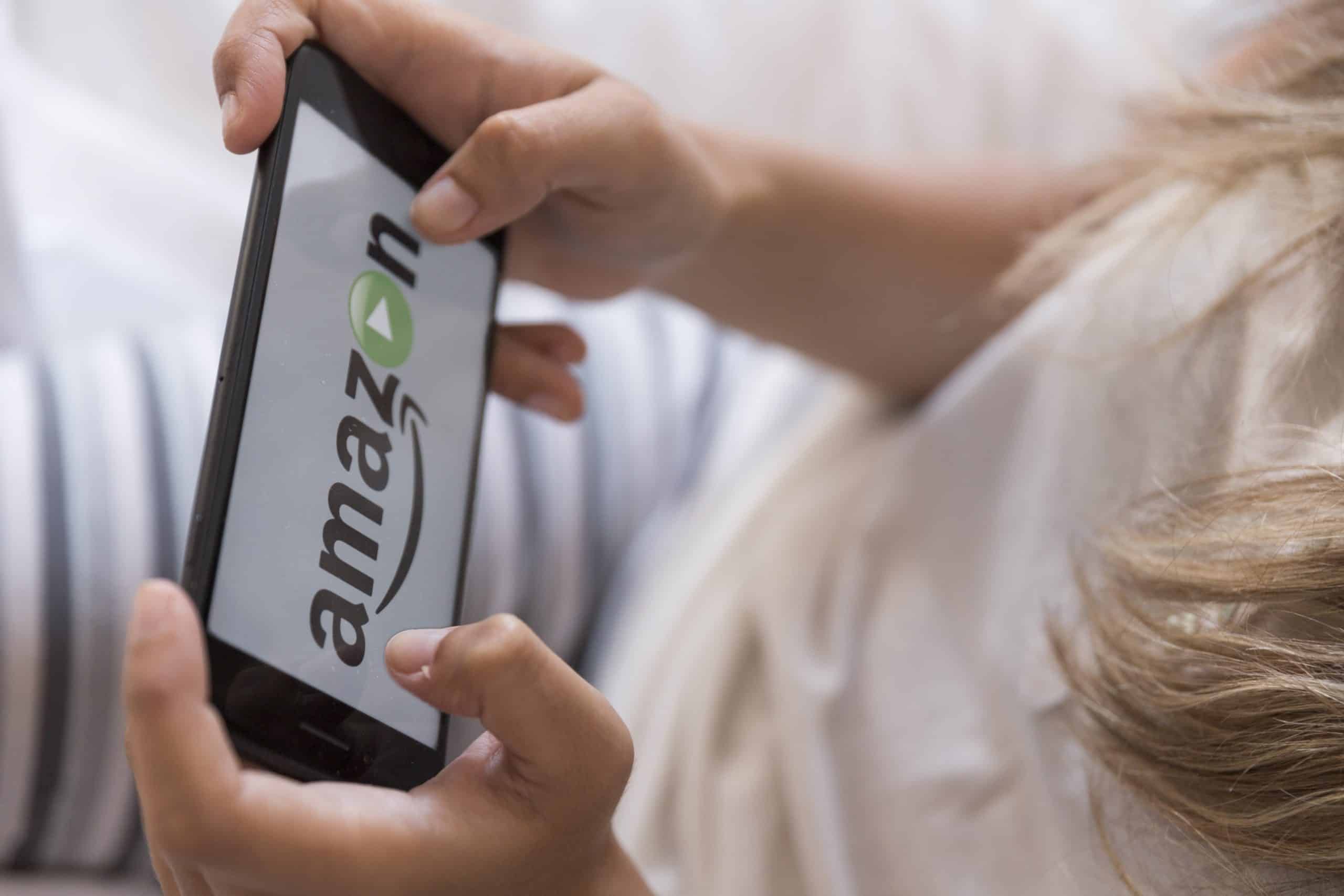 Close-up of a person holding a smartphone displaying the Amazon Prime logo with the play button, likely indicating a streaming service. The person appears to be lying down on a bed, wearing a light-colored shirt and striped pants, ready to rent or buy content for their next movie night.