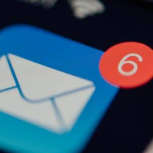 A close-up of an email app icon on an iOS smartphone screen with a red notification badge showing the number 6, indicating there are six unread emails. The image is slightly blurred except for the Apple icon and the notification badge.