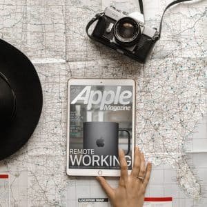 A flat lay photo includes a hat, an old-fashioned camera, and a tablet displaying the cover of AppleMagazine. The magazine cover features a black mug with the Apple logo and the headline "Remote Working." The background is a detailed map with numerous blue pins. #PasaLaPagina