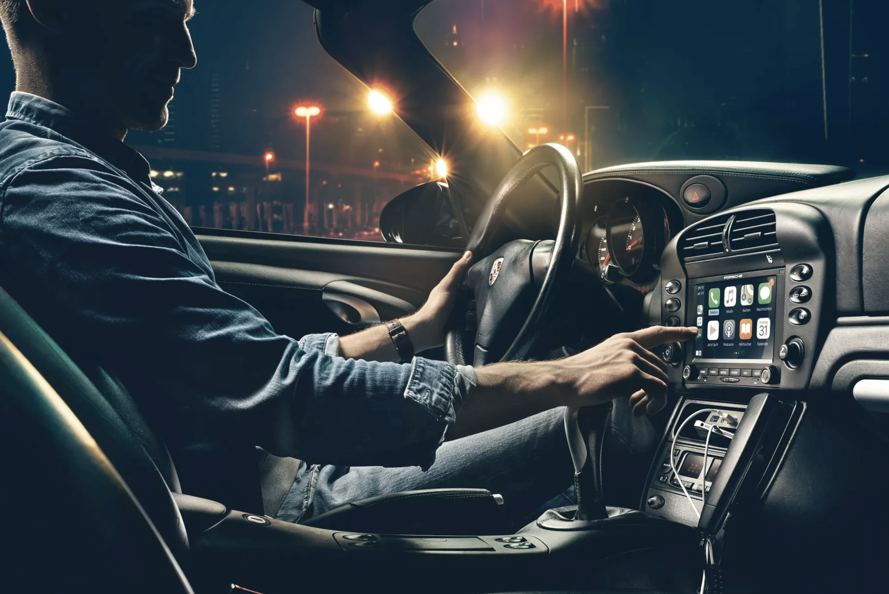 A person wearing a denim shirt is driving a Porsche convertible car at night. They are adjusting the car's Apple CarPlay infotainment system, which displays various app icons on the screen. The car's interior is dimly lit, and city lights are visible through the windshield.