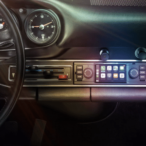 Interior view of a classic Porsche dashboard featuring a mix of vintage controls and a modern touchscreen infotainment system with Apple CarPlay. The steering wheel, speedometer, and other classic gauges are visible, while sunlight creates a gradient effect on the elegant dashboard.