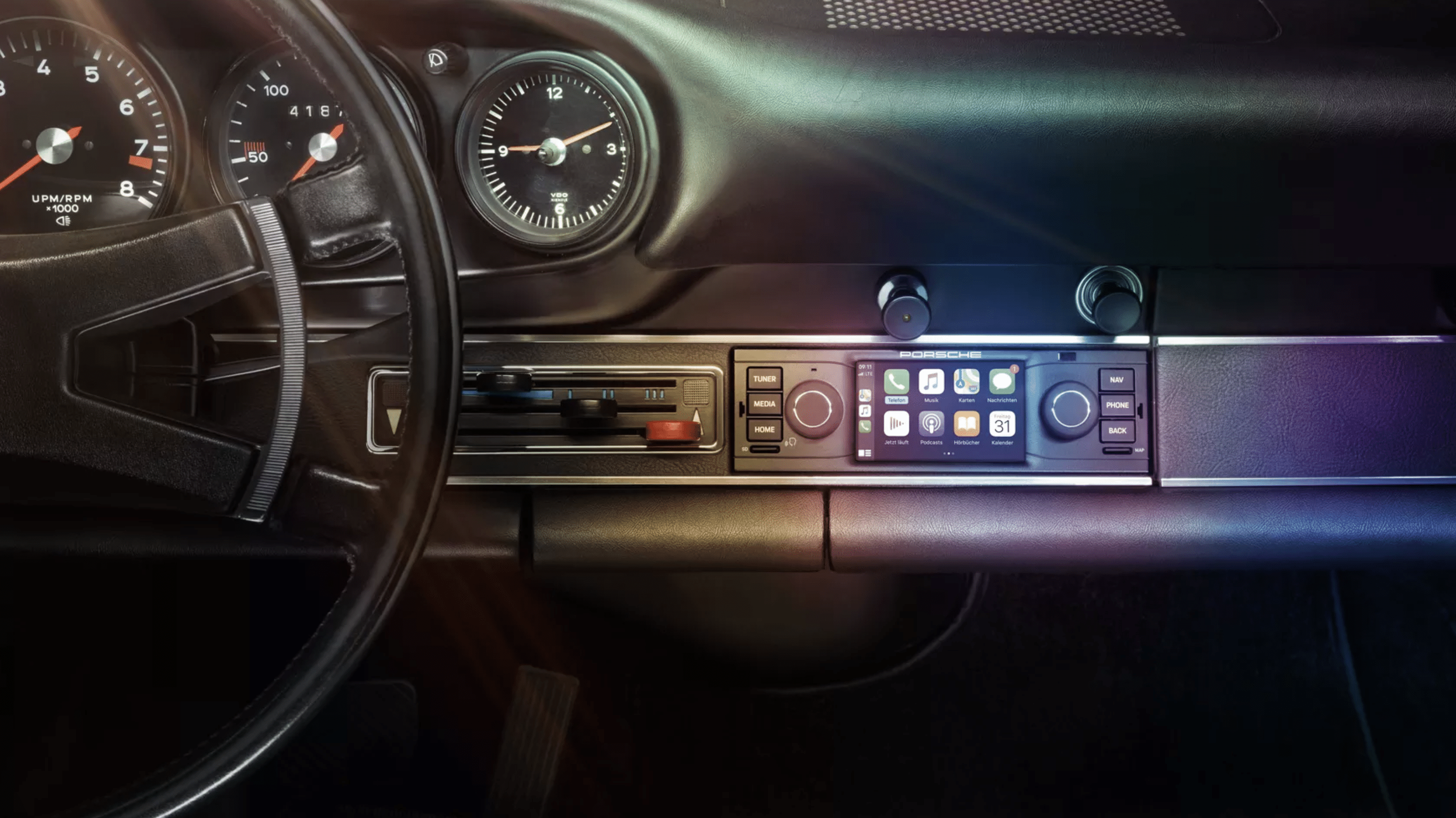 Interior view of a classic Porsche dashboard featuring a mix of vintage controls and a modern touchscreen infotainment system with Apple CarPlay. The steering wheel, speedometer, and other classic gauges are visible, while sunlight creates a gradient effect on the elegant dashboard.