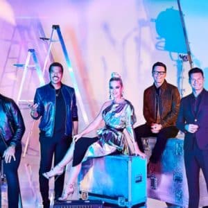 A colorful studio backdrop with five people posing. Three men stand while one woman in a metallic dress sits on a case, and another man stands beside her. There are stage lights, ladders, and guitars in the background, giving an American Idol behind-the-scenes vibe.