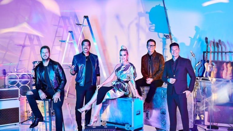 A colorful studio backdrop with five people posing. Three men stand while one woman in a metallic dress sits on a case, and another man stands beside her. There are stage lights, ladders, and guitars in the background, giving an American Idol behind-the-scenes vibe.