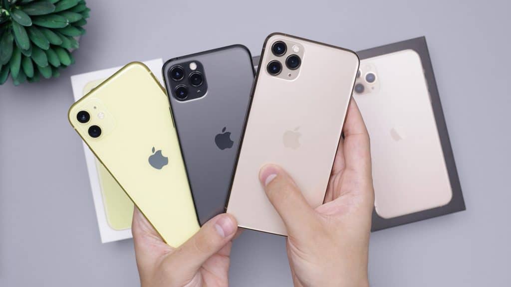 A pair of hands holding four different iPhone models against a gray background. The iPhones are in yellow, black, and two shades of gold. All phones have the Apple logo on the back; two have triple cameras while the others have dual cameras. A plant is visible in the corner, signaling recovery in the smartphone market.