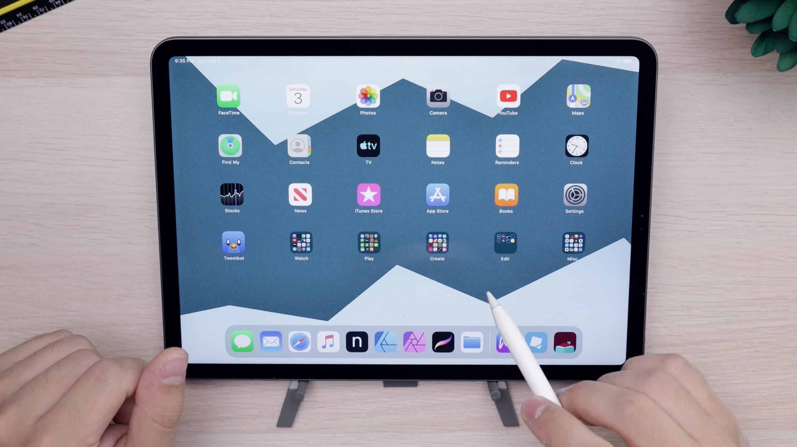 A person holding a stylus and pointing at an iPad Pro screen displaying various app icons, including Safari, Messages, Photos, Camera, YouTube, and others. The tablet boasts a mountain-themed wallpaper. A potted plant is partially visible to the right. The same processor powers this sleek device with new details.