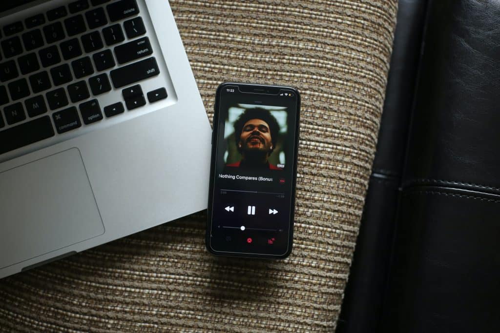 A smartphone displaying a music player app screen rests on a textured surface next to a partially visible open laptop. The music player shows a song by an artist with a bearded man’s face as the cover art and controls for play, rewind, and fast-forward. Nearby, Apple Arcade's logo shines brightly on the screen, hinting at the gaming adventures awaiting in 20 countries.