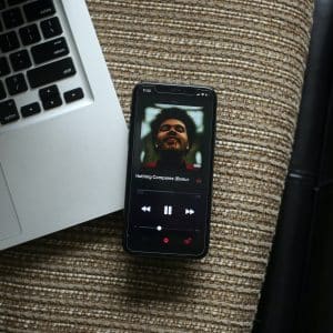 A smartphone displaying a music player app screen rests on a textured surface next to a partially visible open laptop. The music player shows a song by an artist with a bearded man’s face as the cover art and controls for play, rewind, and fast-forward. Nearby, Apple Arcade's logo shines brightly on the screen, hinting at the gaming adventures awaiting in 20 countries.