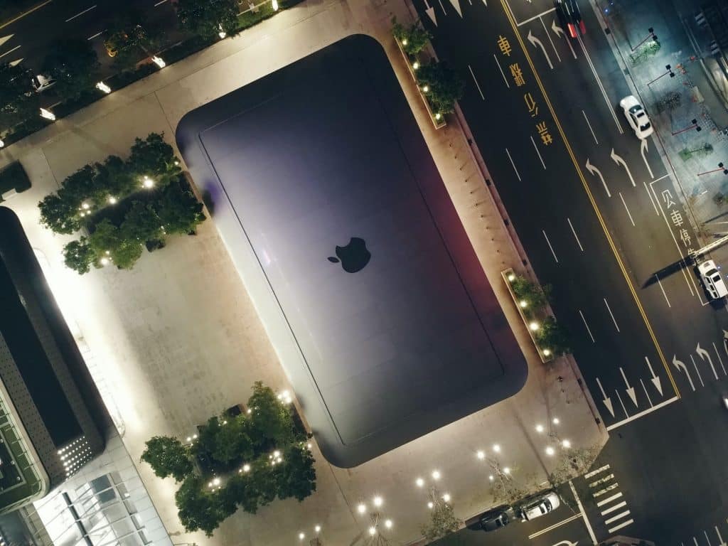 An aerial view of an Apple store at night shows the large glass ceiling with the Apple logo prominently displayed. The surrounding area, lit by streetlights and featuring adjoining trees and roads with little traffic, will look even more inviting when it reopens next month.