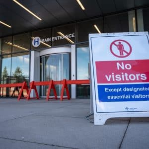 Sign in front of the entrance to a hospital that reads "No visitors except designated essential visitors." The entrance has red barriers and glass doors, with a hospital symbol and "Main Entrance" sign above. This facility also serves as a COVID-19 testing site.