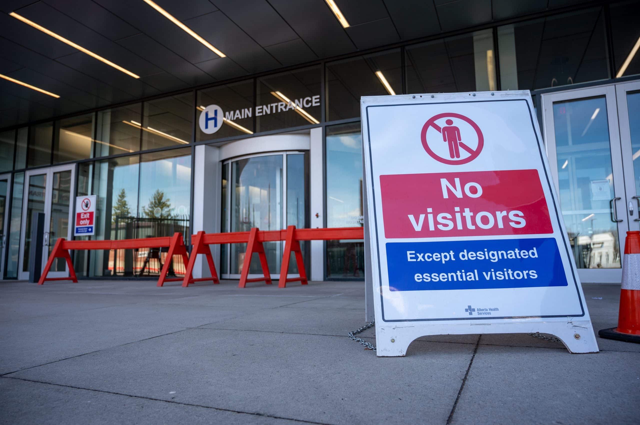 Sign in front of the entrance to a hospital that reads "No visitors except designated essential visitors." The entrance has red barriers and glass doors, with a hospital symbol and "Main Entrance" sign above. This facility also serves as a COVID-19 testing site.