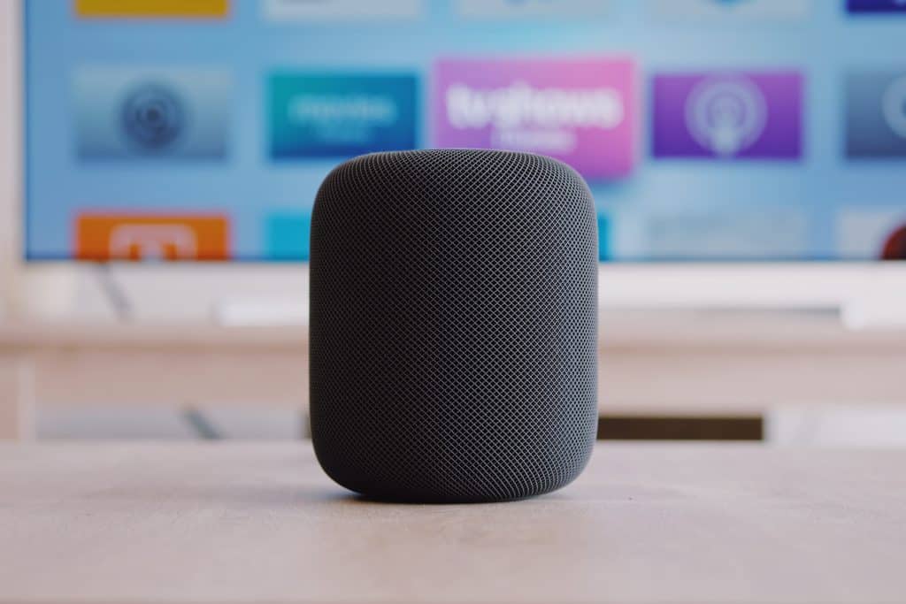 A black, cylindrical HomePod 2 sits on a table in front of a blurred TV screen displaying various colorful app icons. The speaker, with its mesh-like exterior and modern design, boasts features that make it a must-have on any tech wishlist.