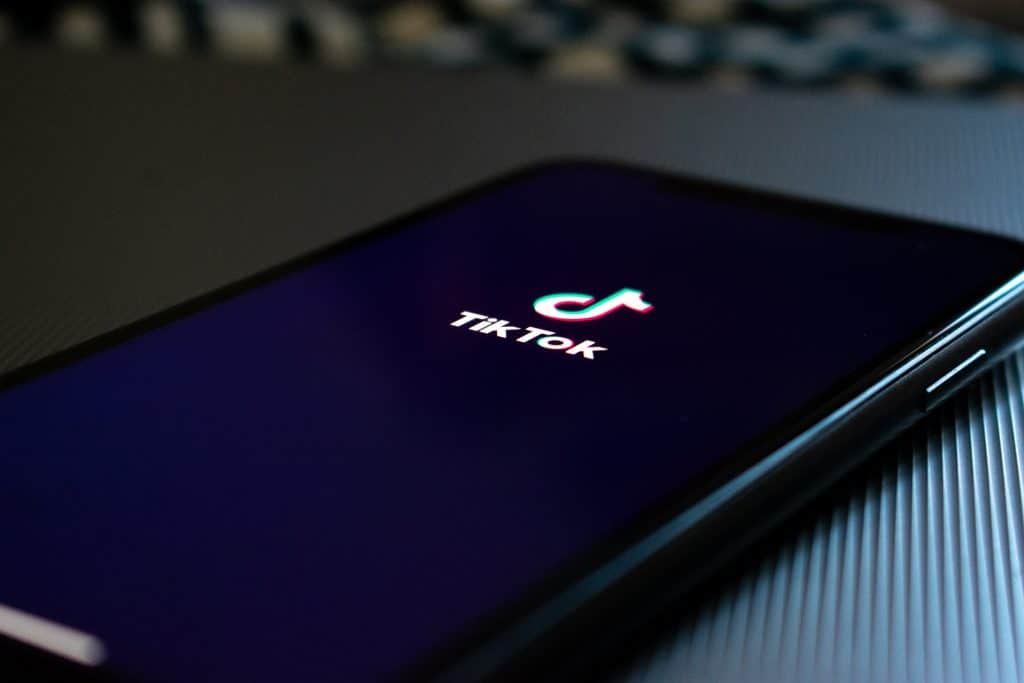 A smartphone screen with the TikTok logo on a black background is displayed. The phone, an Android device, is placed on a dark, textured surface and slightly angled to show its edge. The TikTok logo features a vibrant musical note above the text "TikTok.