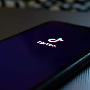 A smartphone screen with the TikTok logo on a black background is displayed. The phone, an Android device, is placed on a dark, textured surface and slightly angled to show its edge. The TikTok logo features a vibrant musical note above the text "TikTok.