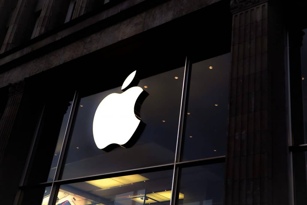 A large illuminated Apple logo is prominently displayed on the glass facade of an Apple Store, flanked by tall, dark pillars. The store's interior is dimly visible, with modern lighting and a glimpse of electronic products inside, reflecting why Apple became the most imitated brand by Q1 2020.