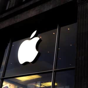 A large illuminated Apple logo is prominently displayed on the glass facade of an Apple Store, flanked by tall, dark pillars. The store's interior is dimly visible, with modern lighting and a glimpse of electronic products inside, reflecting why Apple became the most imitated brand by Q1 2020.