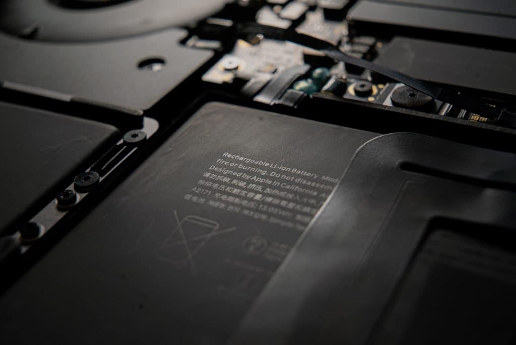 Close-up view of the internal components of a laptop. Focused on a rechargeable lithium battery module, with surrounding circuitry and parts visible. The text on the battery mentions disposal information and its design origin as California. An Apple macOS update includes Battery Health Management features.