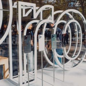 A store window display features three smartphones mounted on stands. The backdrop showcases large metallic models of the numbers "100". Outside, people are gathered on a busy street, with trees and vehicles visible on the road. An Apple sign subtly hints at the tech giant's influence in this US retail scene.