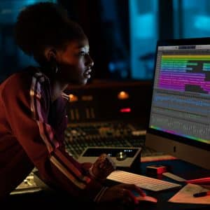 A woman leans over a 23-inch iMac, intensely focusing on its screen displaying colorful audio tracks in a music production software. The room is dimly lit with a blue hue, and a person is seen through the window, possibly recording in a vocal booth. This scene appears to be set in the fourth quarter of 2020.