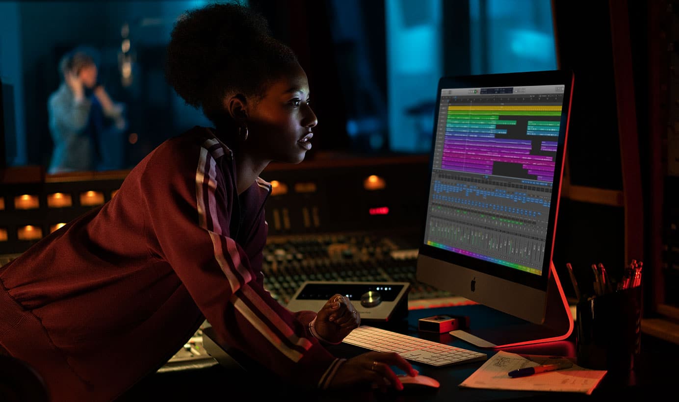 A woman leans over a 23-inch iMac, intensely focusing on its screen displaying colorful audio tracks in a music production software. The room is dimly lit with a blue hue, and a person is seen through the window, possibly recording in a vocal booth. This scene appears to be set in the fourth quarter of 2020.