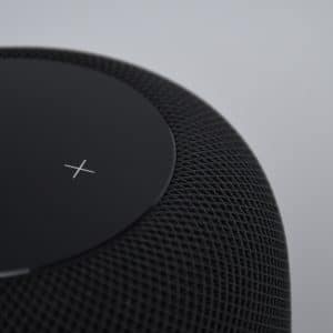 Close-up of a black cylindrical mini HomePod with a mesh exterior. The top surface displays a white 'X' symbol on a black touch-sensitive panel. The background is plain and light-colored.