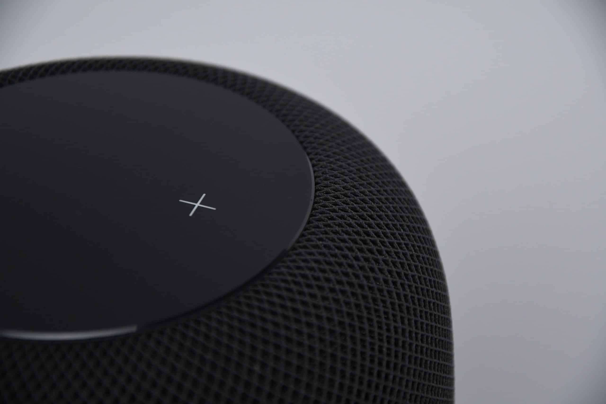 Close-up of a black cylindrical mini HomePod with a mesh exterior. The top surface displays a white 'X' symbol on a black touch-sensitive panel. The background is plain and light-colored.