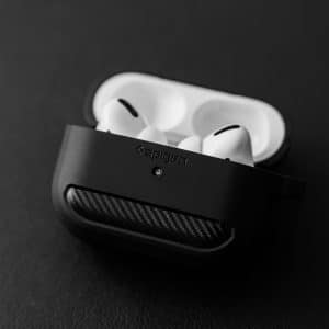 A pair of white wireless earbuds with silicone tips sit in an open black charging case on a dark surface. The charging case has a textured pattern and the brand name "Spigen" is visible on it. These next-generation AirPods are set for an online release soon. The background is out of focus.