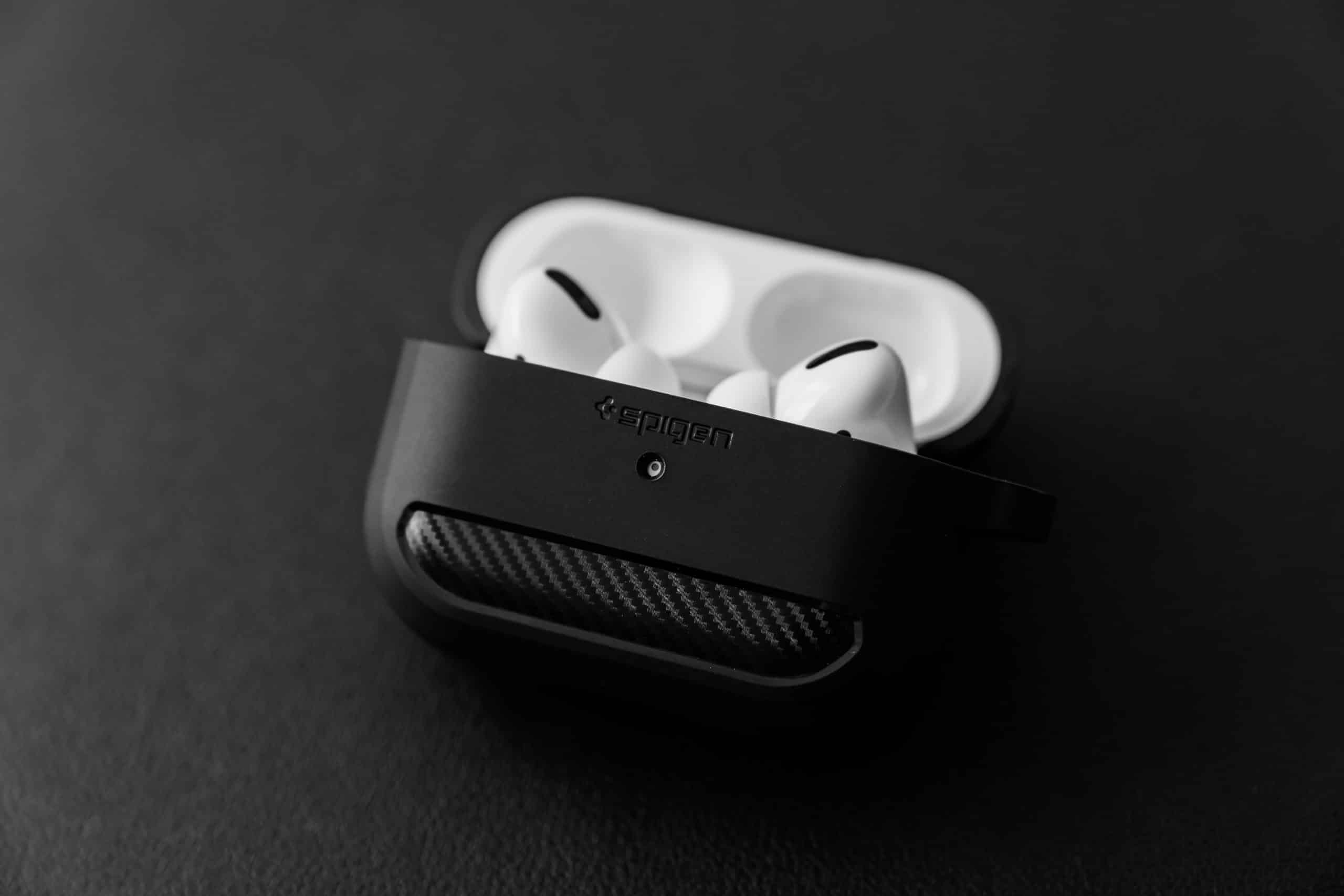 A pair of white wireless earbuds with silicone tips sit in an open black charging case on a dark surface. The charging case has a textured pattern and the brand name "Spigen" is visible on it. These next-generation AirPods are set for an online release soon. The background is out of focus.