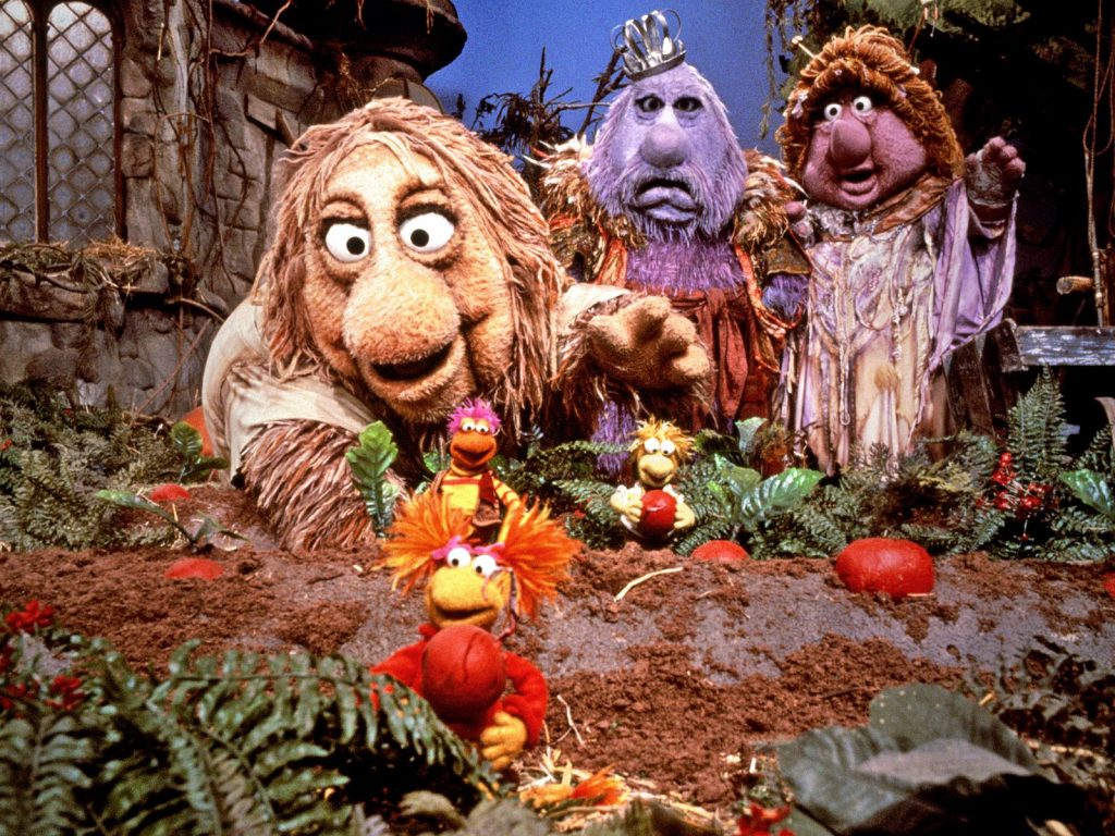 A group of colorful, puppet-like characters stand in a whimsical, forest-like setting reminiscent of Fraggle Rock. In the foreground, smaller creatures with vibrant colors gather among red objects, while larger, shaggy figures loom in the background with expressive faces and fantasy attire. Welcome to Apple TV+.