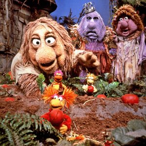 A group of colorful, puppet-like characters stand in a whimsical, forest-like setting reminiscent of Fraggle Rock. In the foreground, smaller creatures with vibrant colors gather among red objects, while larger, shaggy figures loom in the background with expressive faces and fantasy attire. Welcome to Apple TV+.