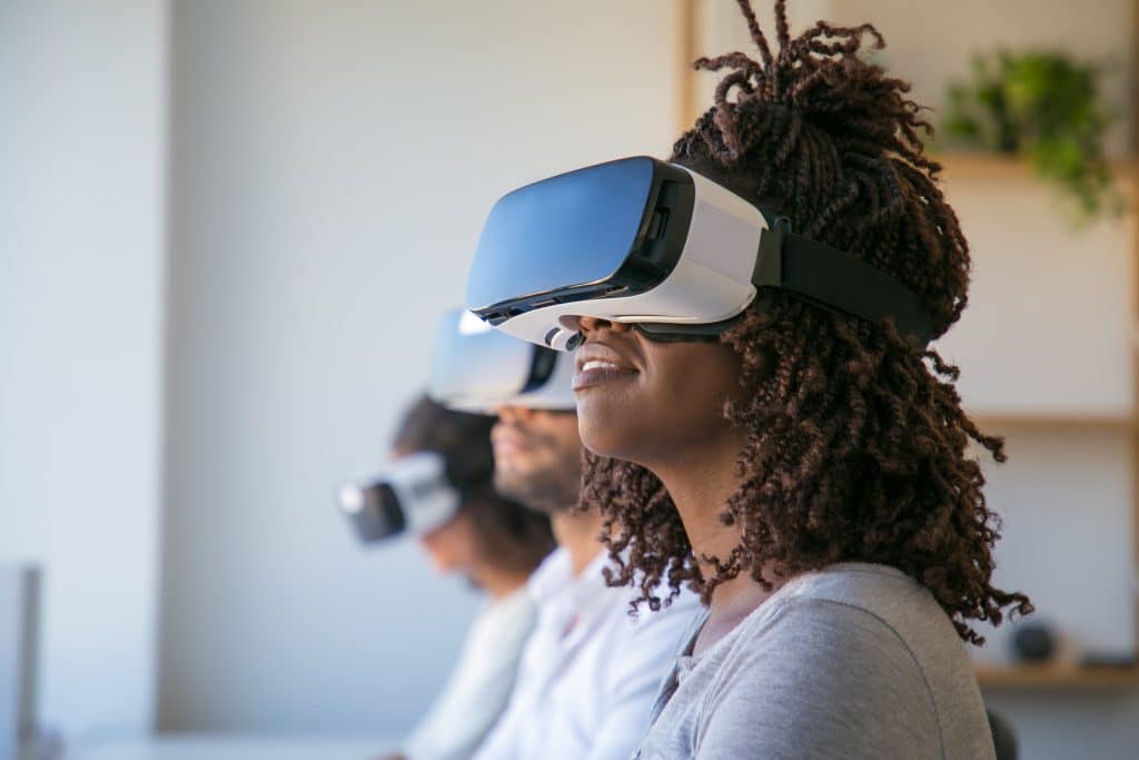 Three people are seated, wearing virtual reality headsets. The person in the foreground has curly hair and is smiling slightly, while the two others, slightly out of focus, are in the background. The setting seems to be an indoor space with shelves on the wall and custom QR codes for quick access to Apple iOS 14 features.