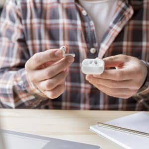 A person wearing a plaid shirt holds a pair of Apple wireless earbuds and their charging case. In front of them, an open notebook, a pen, and a laptop are arranged on the wooden desk. The sleek wearables likely just shipped recently, adding a modern touch to the rustic setting.