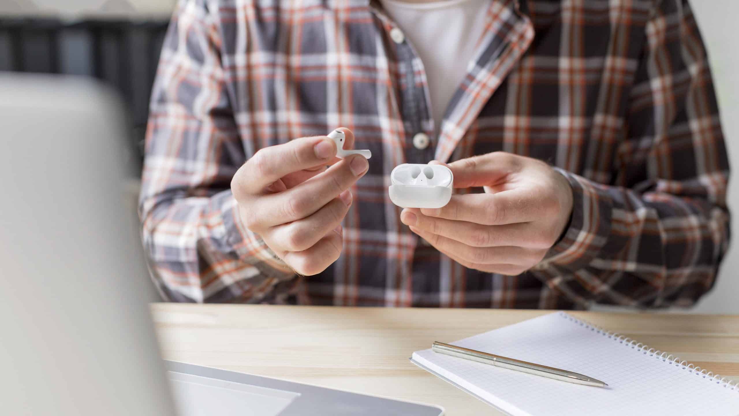 A person wearing a plaid shirt holds a pair of Apple wireless earbuds and their charging case. In front of them, an open notebook, a pen, and a laptop are arranged on the wooden desk. The sleek wearables likely just shipped recently, adding a modern touch to the rustic setting.