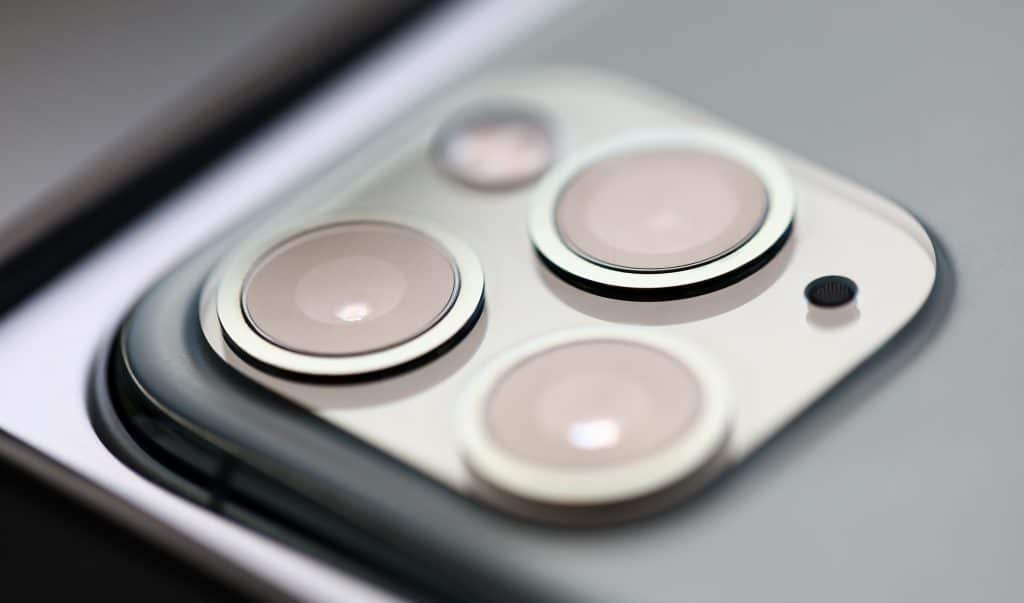 Close-up view of a smartphone's camera module featuring three lenses and a small sensor, set in a metallic square bump on the back of the device, showcasing Apple's sleek production quality with assembly taking place in India.