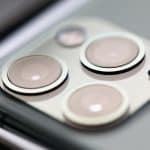 Close-up view of a smartphone's camera module featuring three lenses and a small sensor, set in a metallic square bump on the back of the device, showcasing Apple's sleek production quality with assembly taking place in India.