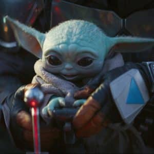 A small green creature with large eyes and long ears, wearing a beige robe, is held by a person in metallic armor. The creature, known as "Baby Yoda," sits serenely in front of control levers. Streaming around the world on Disney+, this iconic scene has entranced countless subscribers.