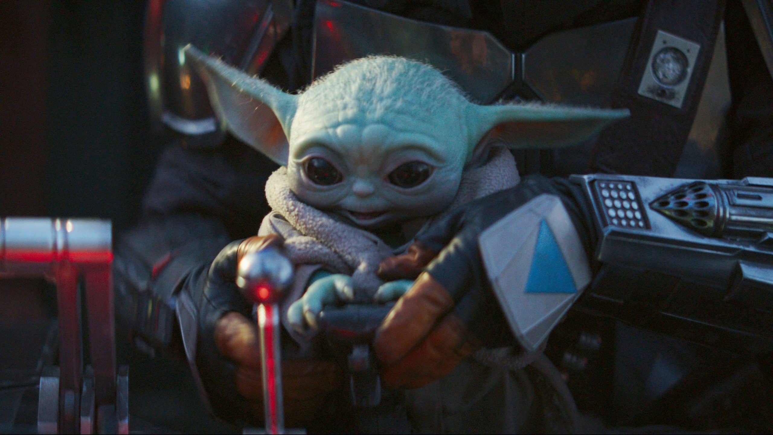 A small green creature with large eyes and long ears, wearing a beige robe, is held by a person in metallic armor. The creature, known as "Baby Yoda," sits serenely in front of control levers. Streaming around the world on Disney+, this iconic scene has entranced countless subscribers.