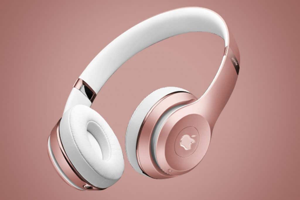 A pair of over-ear headphones with metallic rose gold and white accents. The headband and ear cushions are white, while the outer parts of the ear cups and parts of the headband are rose gold. Reminiscent of Apple's new AirPods Studio, the background features a gradient of pink hues.