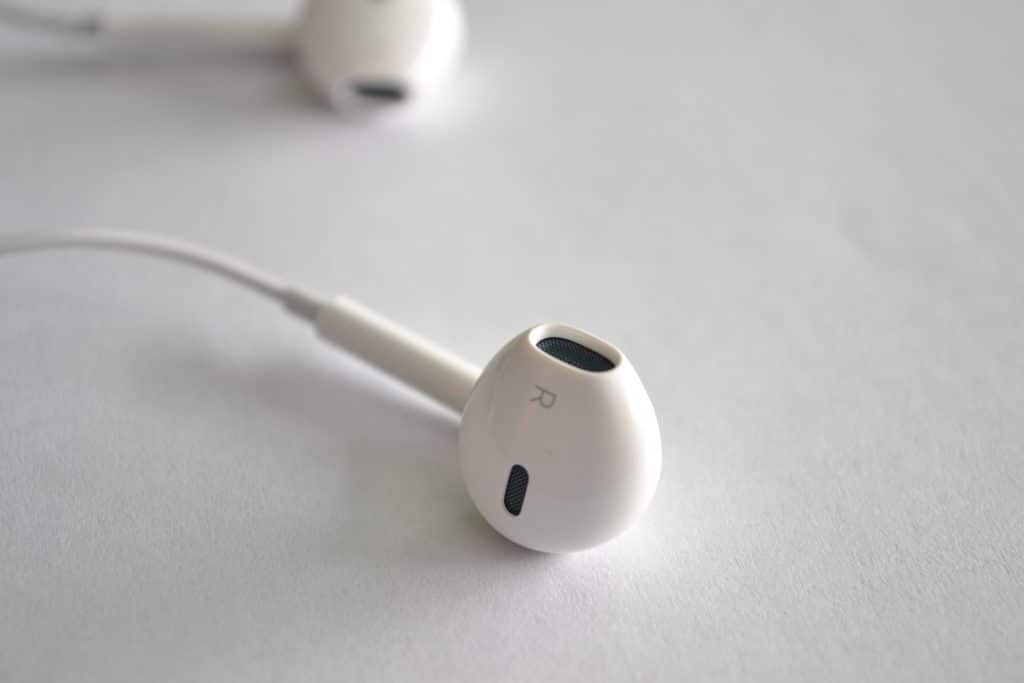 Close-up image of a white Apple EarPod marked with an "R" to indicate it is for the right ear, designed specifically for iPhones. Another EarPod is slightly out of focus in the background, both resting on a clean white surface.