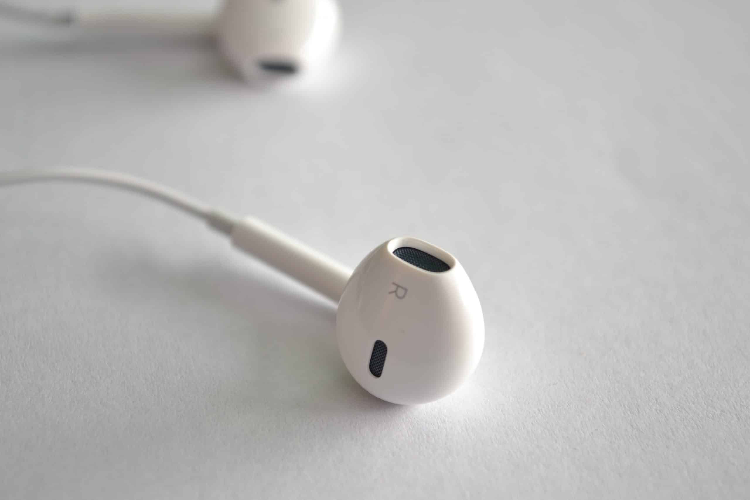 Close-up image of a white Apple EarPod marked with an "R" to indicate it is for the right ear, designed specifically for iPhones. Another EarPod is slightly out of focus in the background, both resting on a clean white surface.