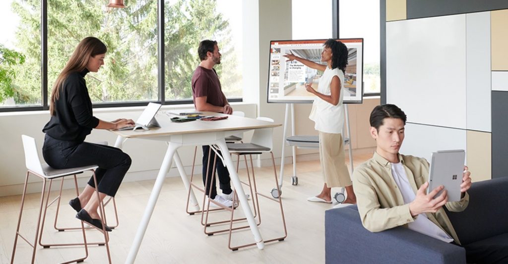 A woman points at a large touchscreen display while standing next to a man. Another woman sits at a high table working on a laptop, likely preparing to pass the Microsoft 70-480 exam. A man sits on a couch using a tablet. They are in a modern, bright office space with large windows and natural light.