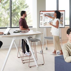 A woman points at a large touchscreen display while standing next to a man. Another woman sits at a high table working on a laptop, likely preparing to pass the Microsoft 70-480 exam. A man sits on a couch using a tablet. They are in a modern, bright office space with large windows and natural light.