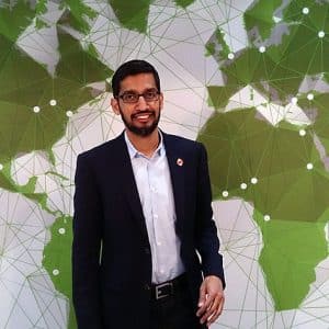 A man wearing a dark suit and glasses stands smiling in front of a green and white world map with a geometric design. The map, reminiscent of Sundar Pichai's vision for the future, appears to be connected with lines and dots, indicating a network or global communication theme.
