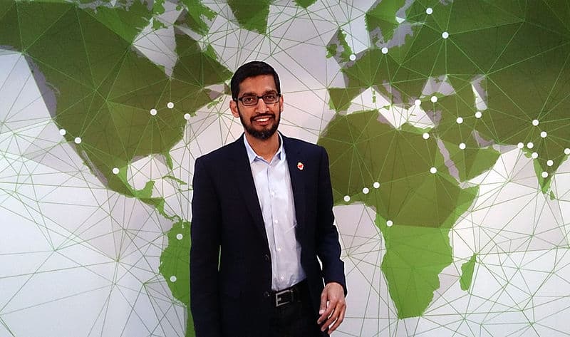 A man wearing a dark suit and glasses stands smiling in front of a green and white world map with a geometric design. The map, reminiscent of Sundar Pichai's vision for the future, appears to be connected with lines and dots, indicating a network or global communication theme.