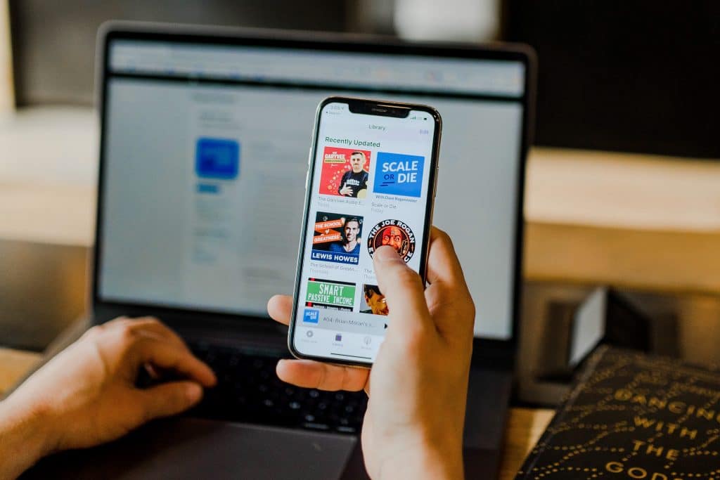 A person is holding a smartphone in one hand, displaying exclusive podcast covers. Behind the smartphone, there's an open laptop on a wooden desk with blurred content on its screen. A notebook sits to the right of the laptop, suggesting a setup ready to compete with Spotify or Apple Podcasts.