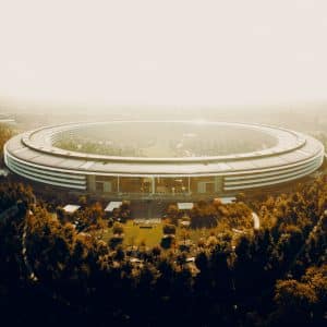 Aerial view of a large, circular, futuristic building surrounded by trees and greenery. The Apple facility has a sleek, modern design with an open interior courtyard and is bathed in warm, golden light, suggesting it is either dawn or dusk. Perfect for staff looking forward to their return to work.