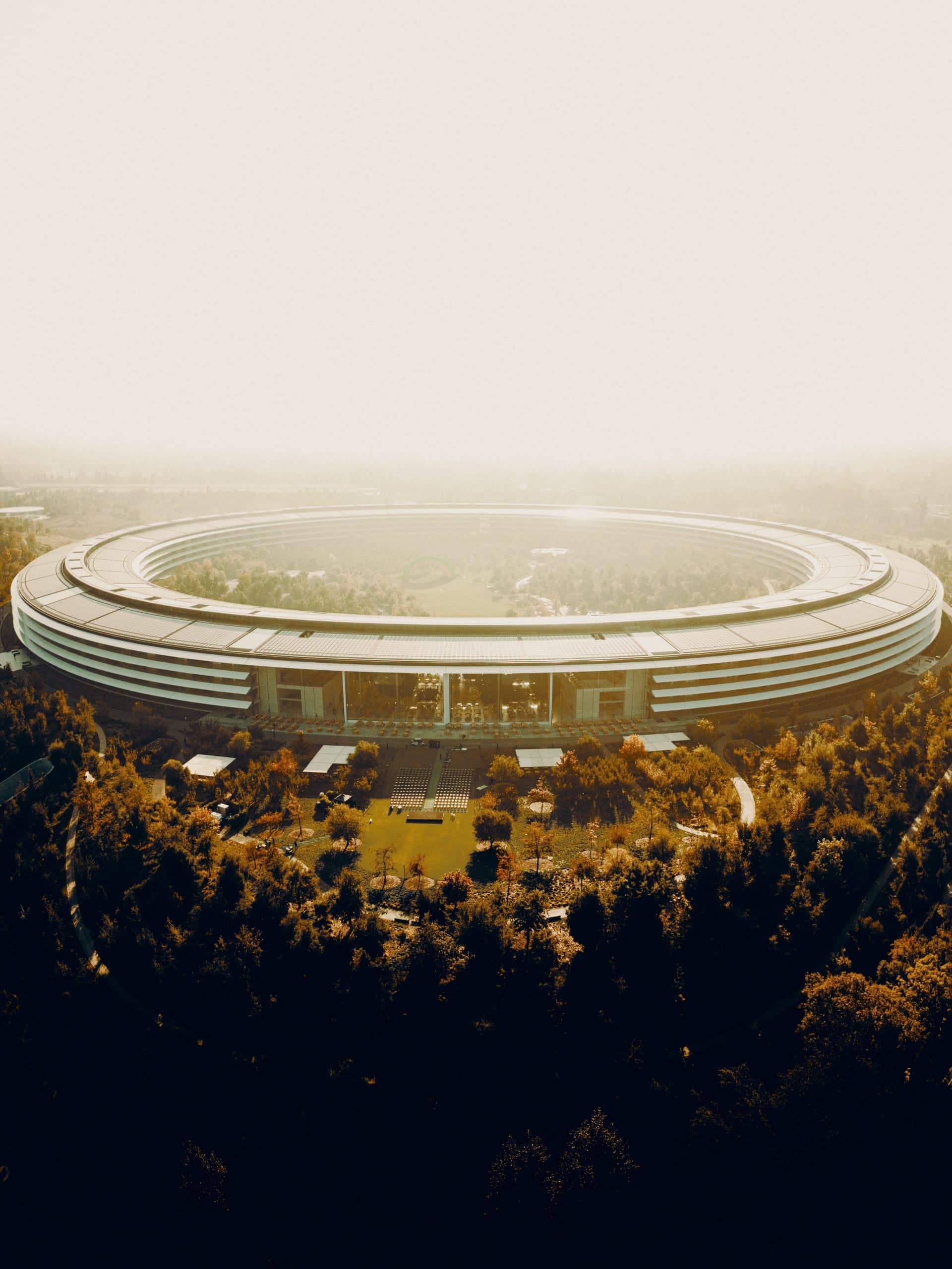 Aerial view of a large, circular, futuristic building surrounded by trees and greenery. The Apple facility has a sleek, modern design with an open interior courtyard and is bathed in warm, golden light, suggesting it is either dawn or dusk. Perfect for staff looking forward to their return to work.