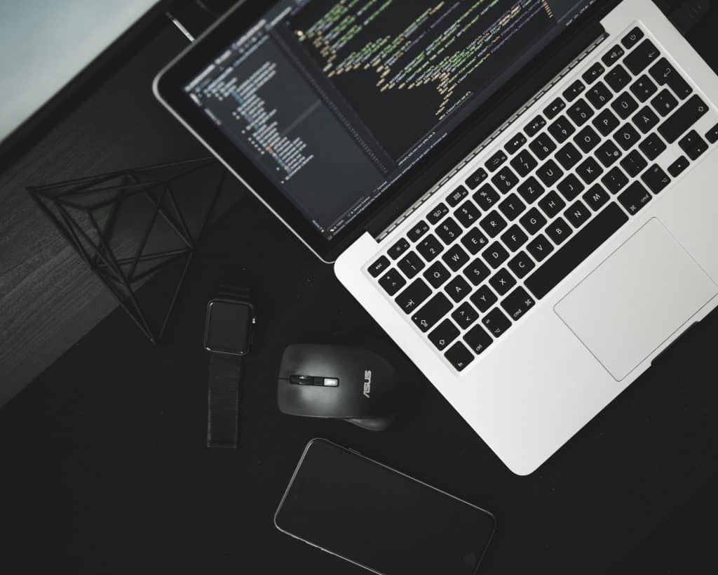 A workspace featuring a laptop with code on the screen, a wireless mouse, a smartwatch, a smartphone, and a geometric decor piece. The setting suggests a tech or programmer's desk with a minimalist and sleek design—a setup growing in popularity alongside the Mac sales increase driven by the coronavirus pandemic.